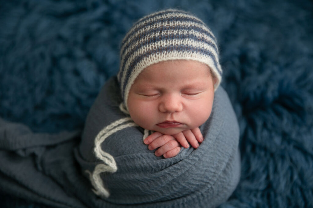 Sleeping newborn baby wrapped in blue fabric wearing knit cap during professional newborn photography session