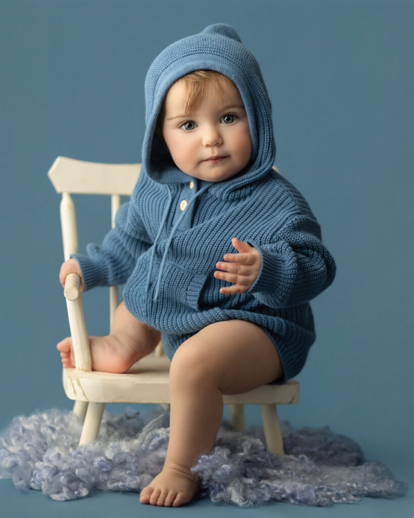 One year old baby in a blue knit outfit sitting on a chair during a milestone photography session at Alternate Angles Photography in Norwood MA