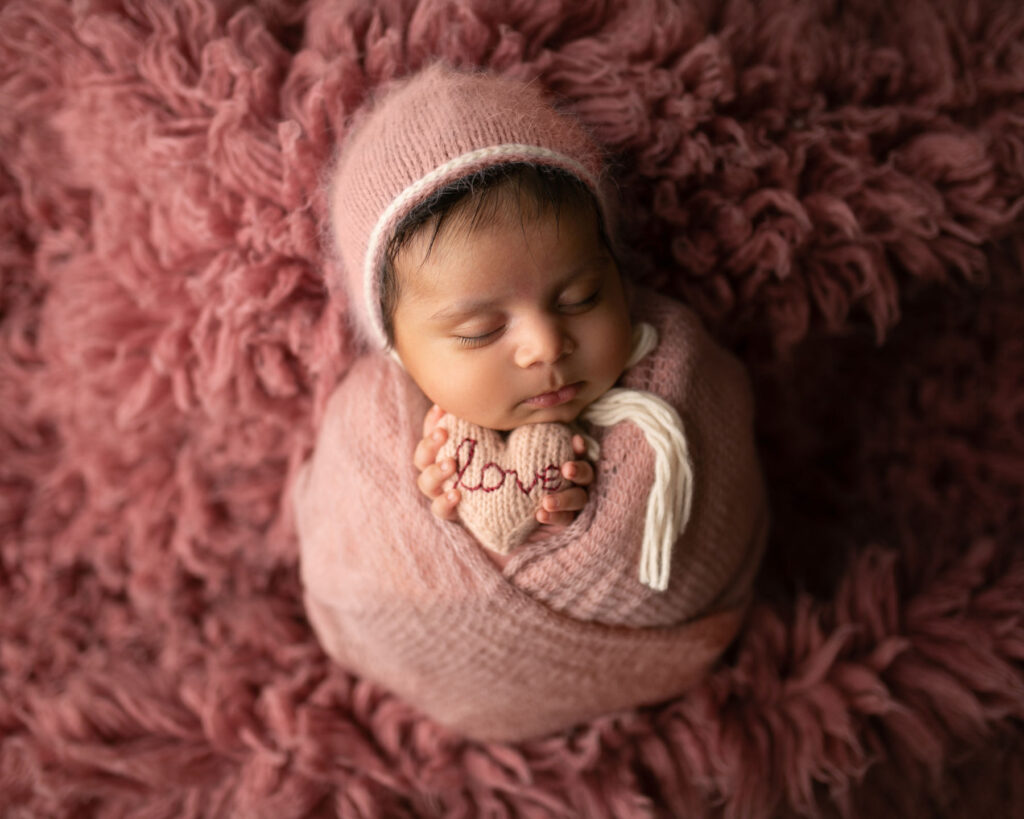 Sleeping newborn baby wrapped in pink knit fabric holding knitted heart with love stitched on front