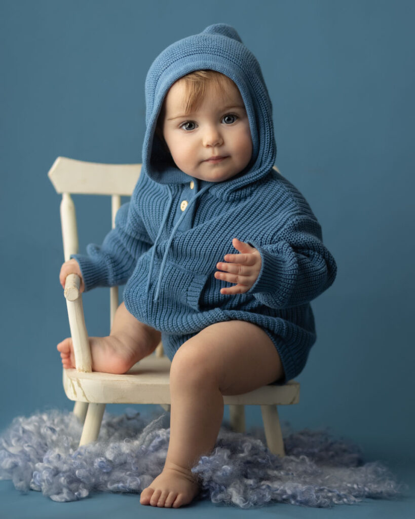 Baby sitting on chair wearing blue hoodie during milestone portrait session.