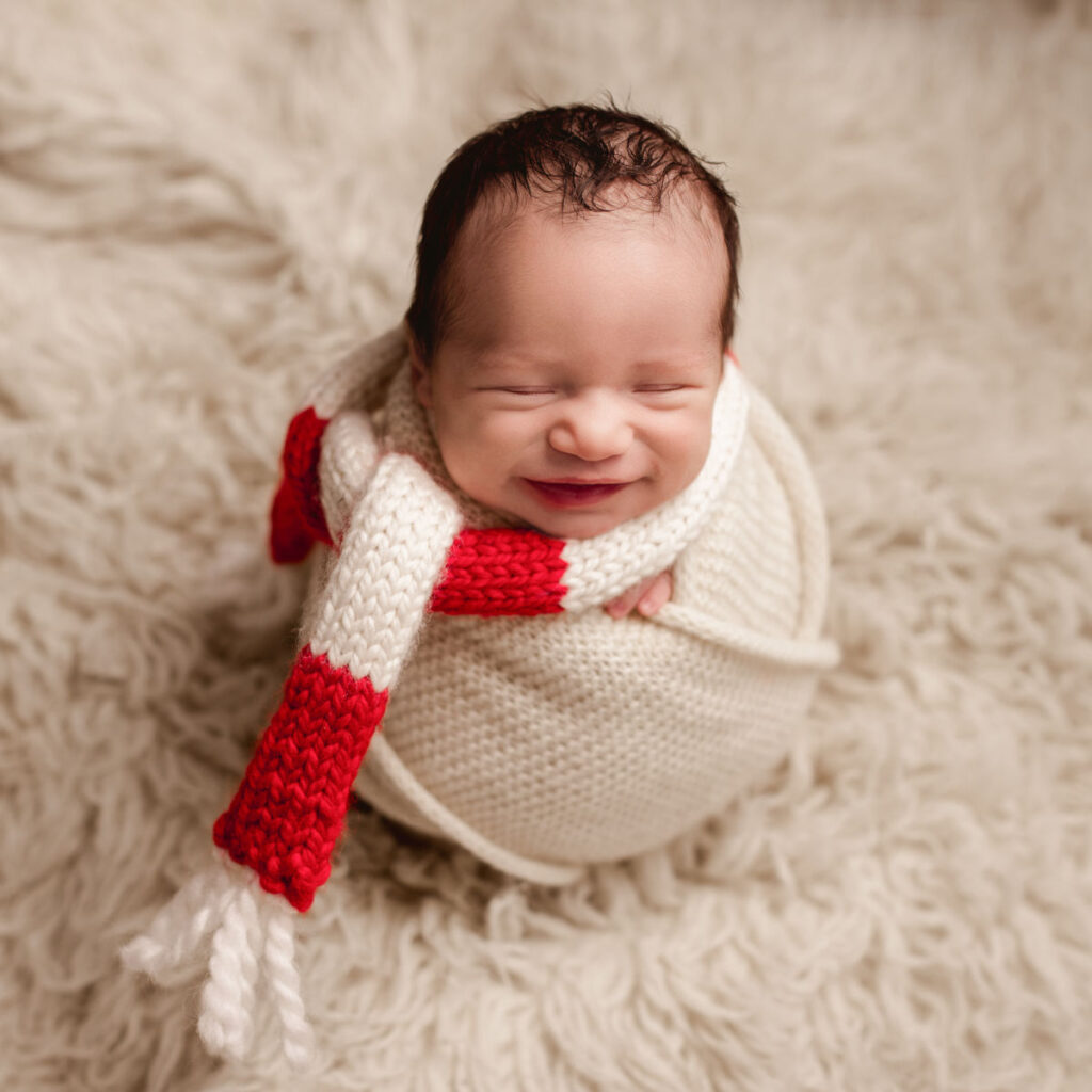 Smiling newborn baby wrapped in a cream knit blanket with a red and white scarf during a professional newborn photography session at Alternate Angles Photography in Norwood MA
