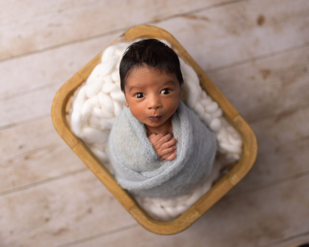Wide-eyed newborn baby boy wrapped in a soft blue blanket and posed in a wooden basket during a newborn photography session