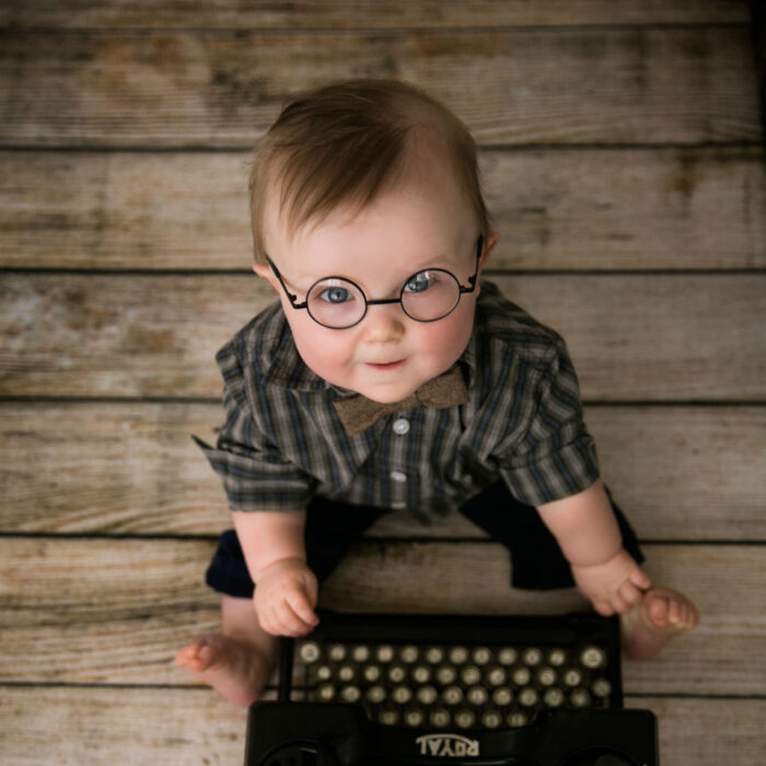 Baby boy wearing glasses sitting beside vintage typewriter during milestone sitter photography session
