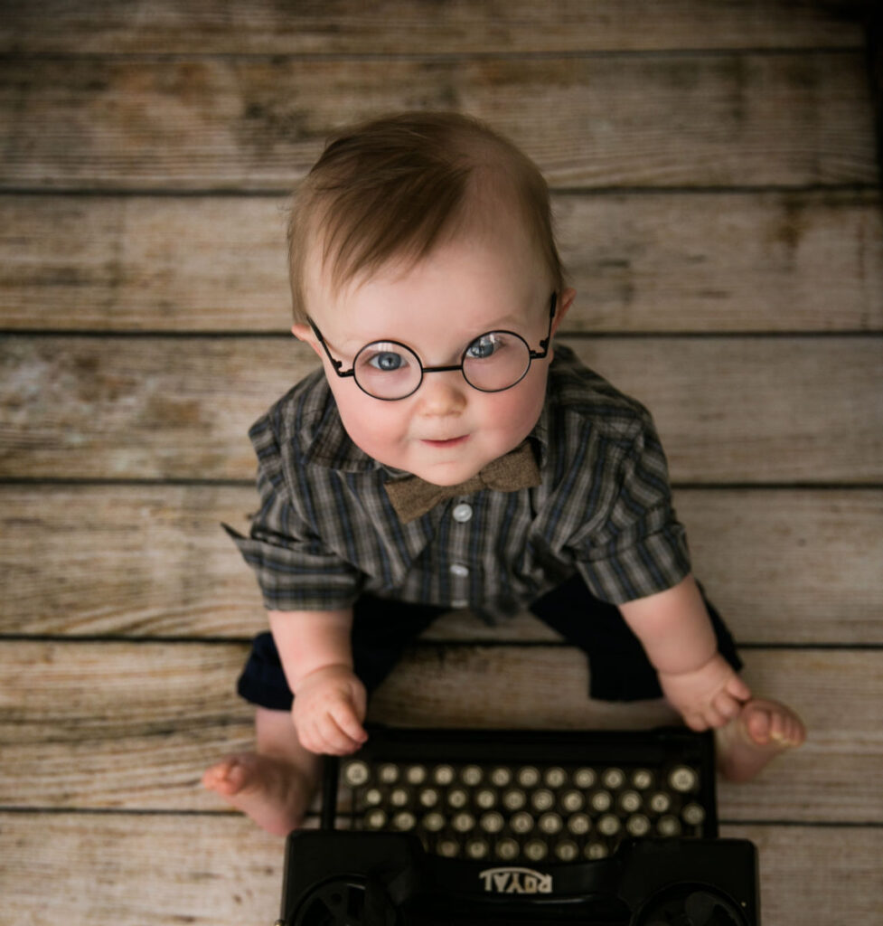 Baby boy wearing glasses sitting beside vintage typewriter during milestone sitter photography session