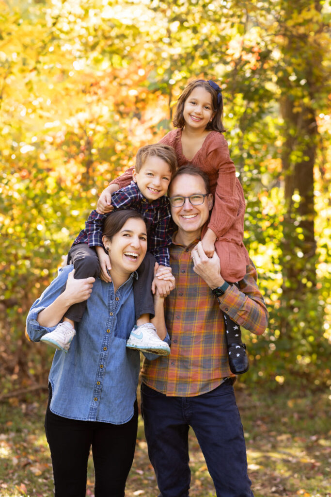 Family portrait session in New England park during fall with smiling parents carrying children among colorful autumn foliage