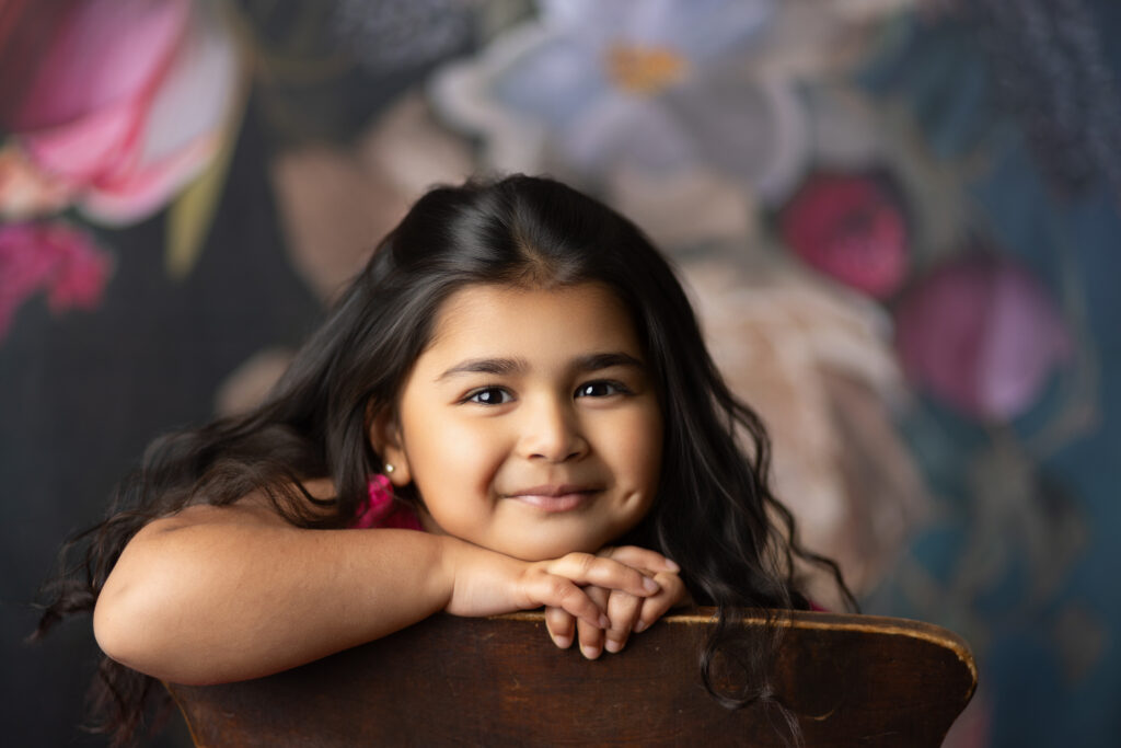 Smiling 4-year-old girl with long dark hair leaning on a wooden chair during a studio milestone portrait session.