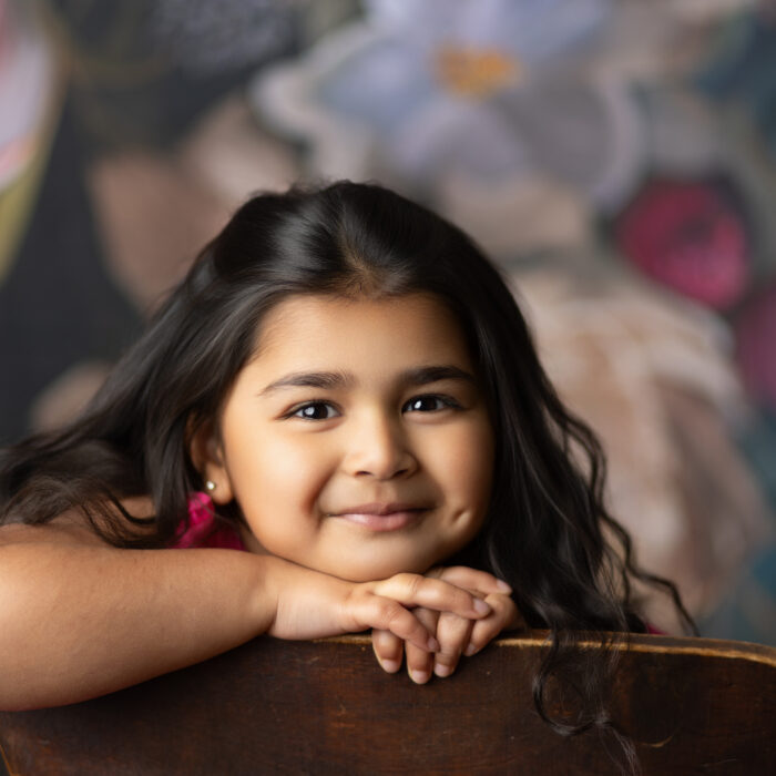 Smiling 4-year-old girl with long dark hair leaning on a wooden chair during a studio milestone portrait session.
