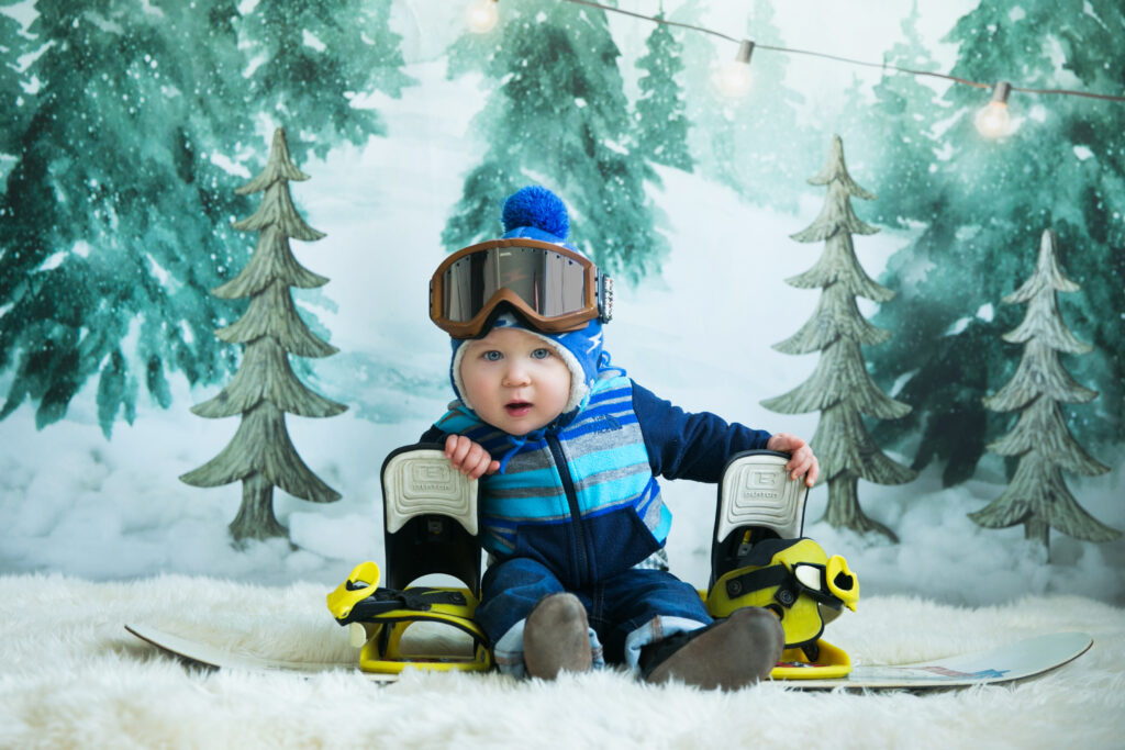 Baby dressed in winter gear sitting on a snowboard during a themed milestone photography session in Norwood, MA