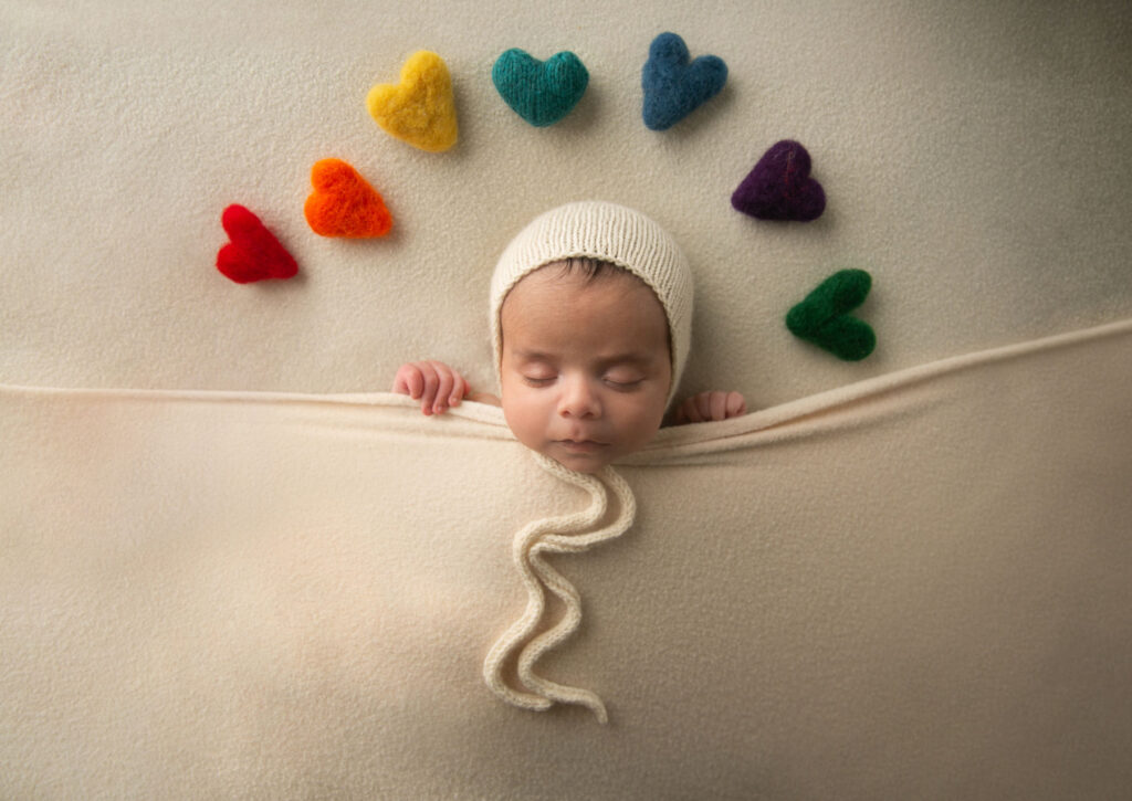 Sleeping newborn baby wrapped in a cream blanket beneath colorful heart decorations during a newborn photography session