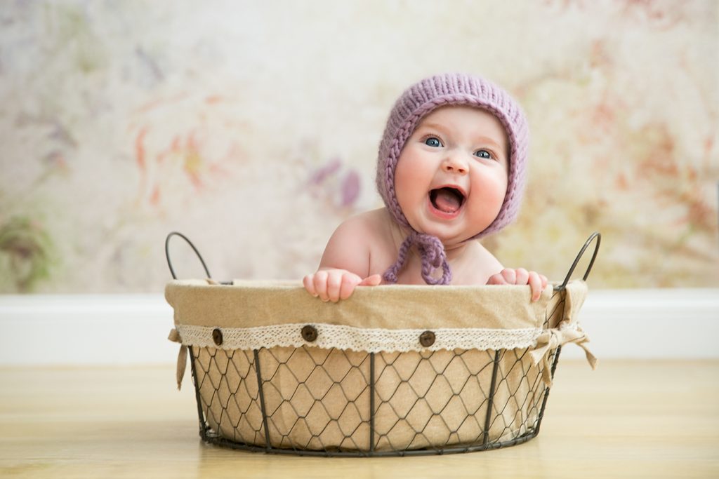 Laughing baby wearing a lavender knitted bonnet while sitting in a rustic basket during a studio milestone session.