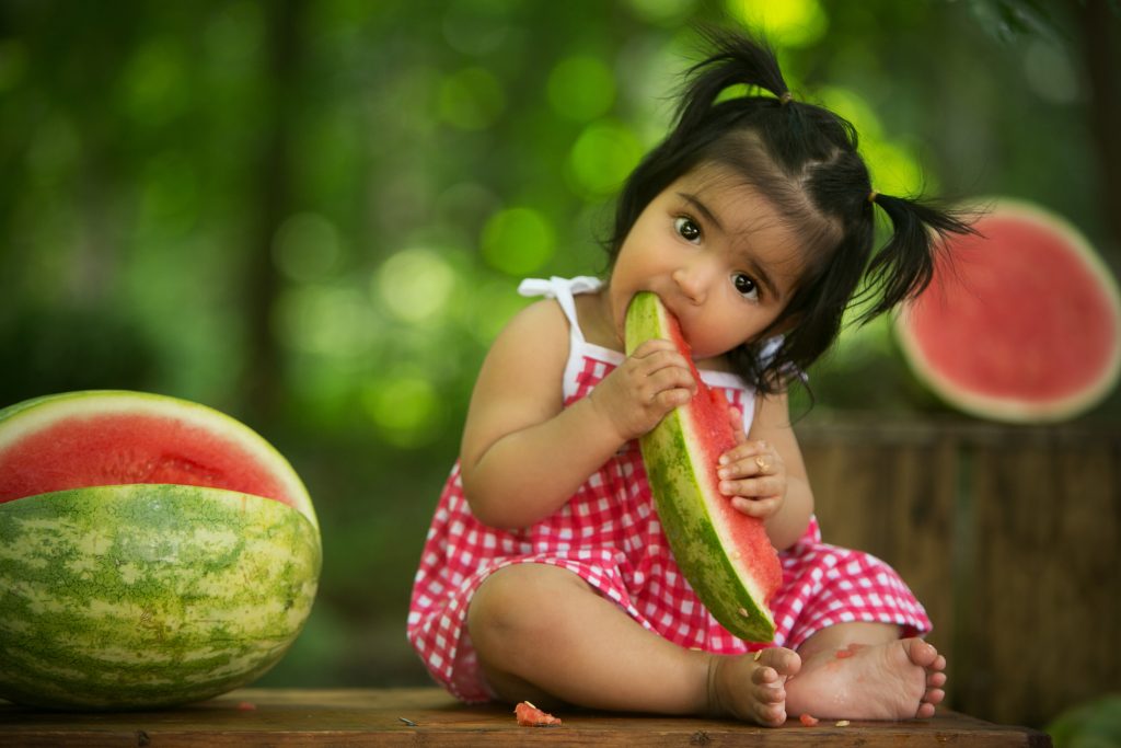 Baby girl in a red gingham outfit eating a watermelon slice during an outdoor summer milestone photography session.