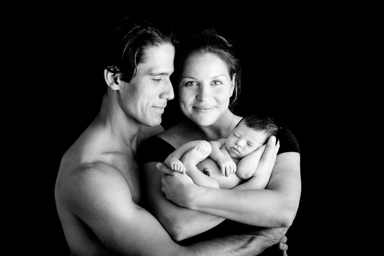 Black-and-white portrait of two parents holding their sleeping newborn baby close against a dark studio background.