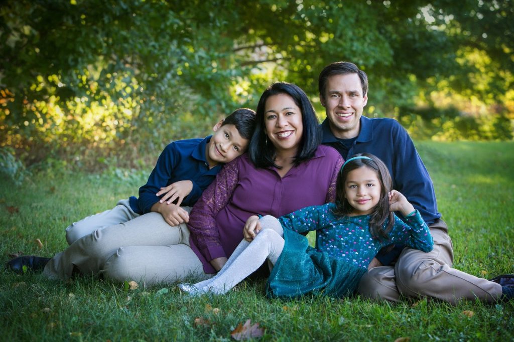 Family of four smiling together while seated on the grass during an outdoor Greater Boston photography session.
