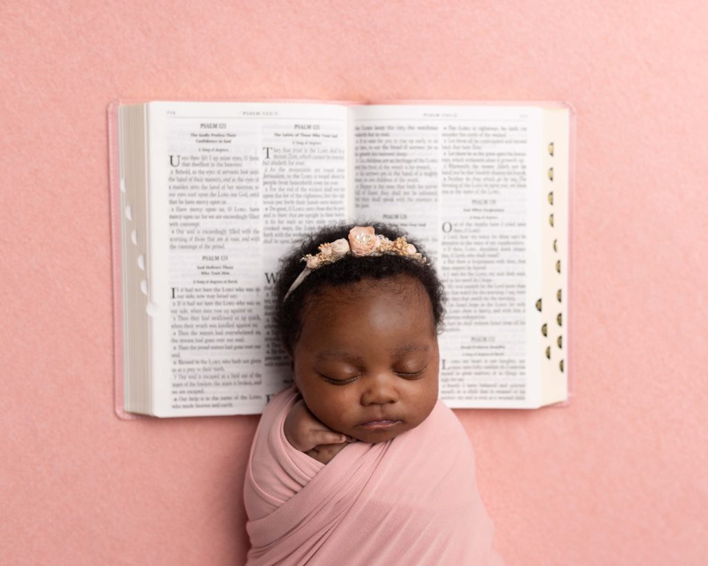 Sleeping newborn girl wrapped in pink and posed beneath an open family Bible during a Norwood newborn photography session.
