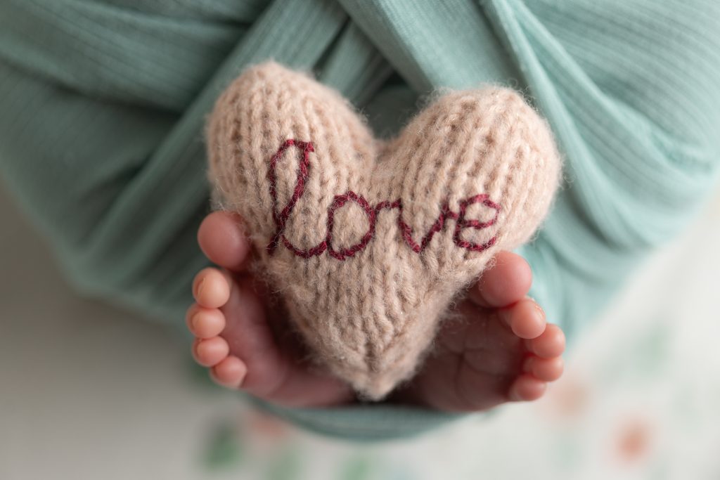 Close-up of newborn feet holding a knitted heart embroidered with the word love.