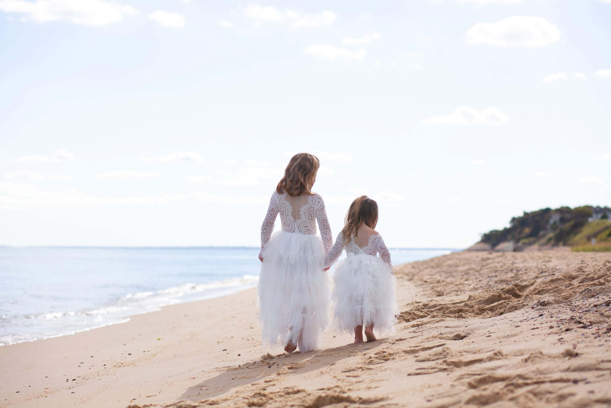Two sisters in white dresses holding hands while walking together along a Massachusetts beach.