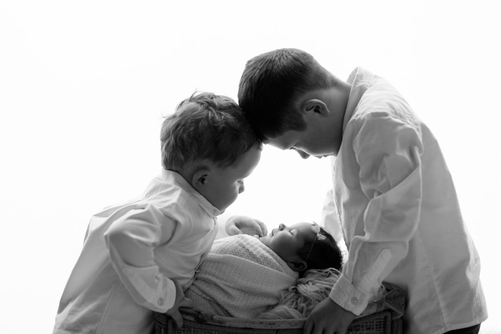 Black and white portrait of two brothers leaning over their newborn baby sister during a professional newborn session.