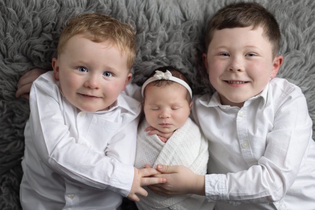 Two proud brothers cuddling their newborn baby sister during a professional newborn photography session in the Boston area.
