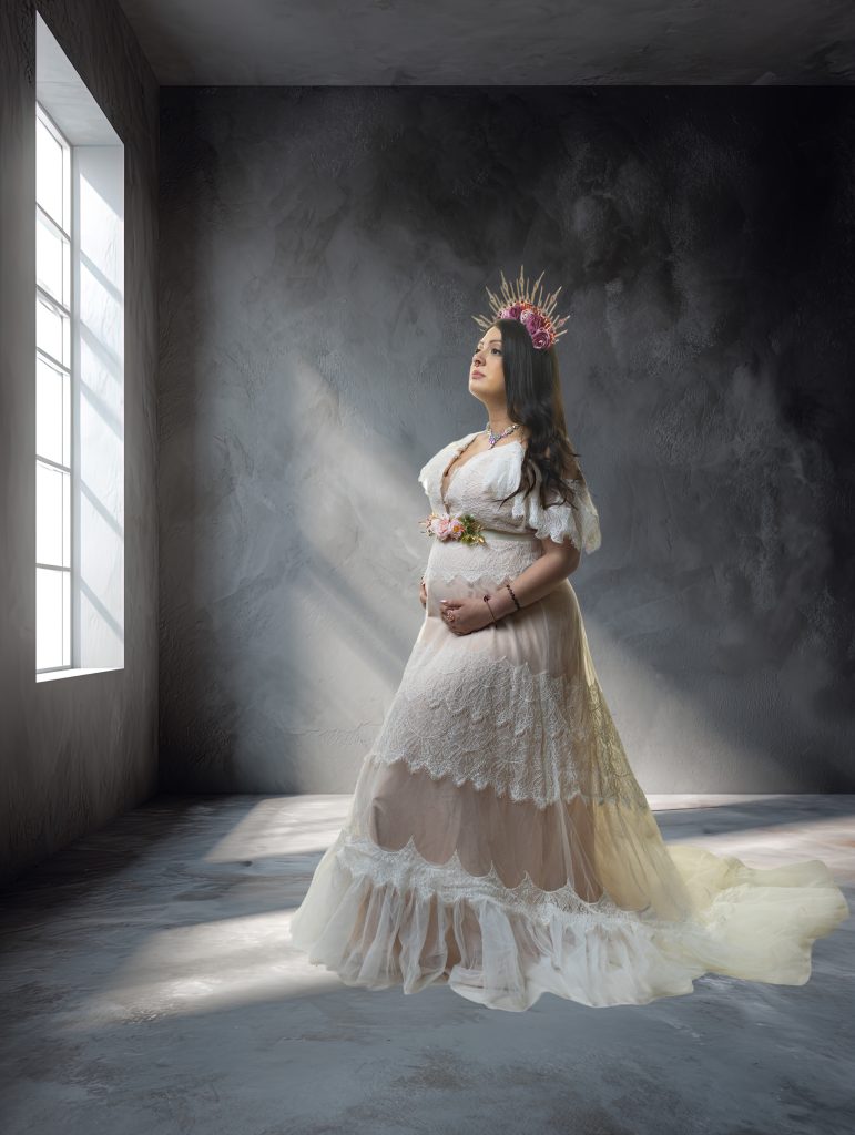 Fine art maternity portrait in a flowing lace gown with dramatic window light in a Boston studio.