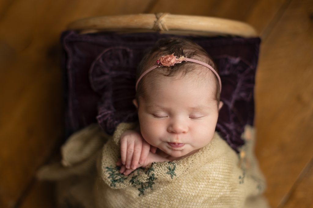 Sleeping newborn baby girl wrapped in a flowered tan blanket with a pink floral headband resting in a basket with purple accents during her Boston newborn session.
