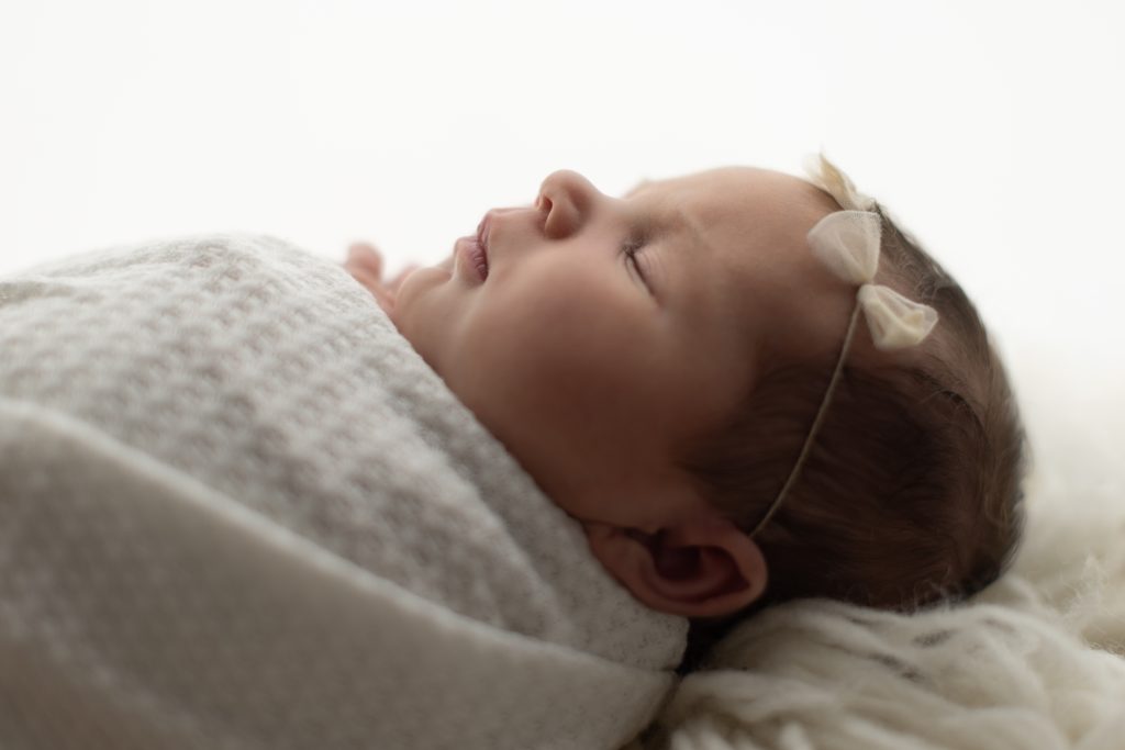 Side profile of a sleeping newborn baby girl wrapped in white with delicate cream bows photographed by a Boston newborn photographer.