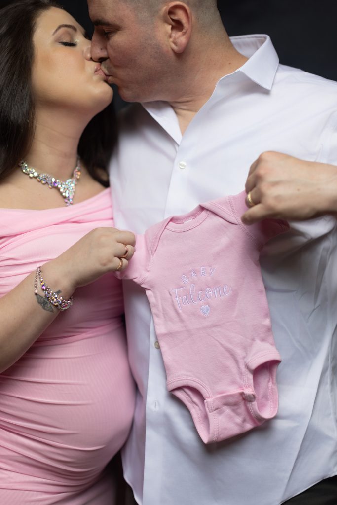 Couple kissing while holding a personalized baby onesie during a Boston maternity session.