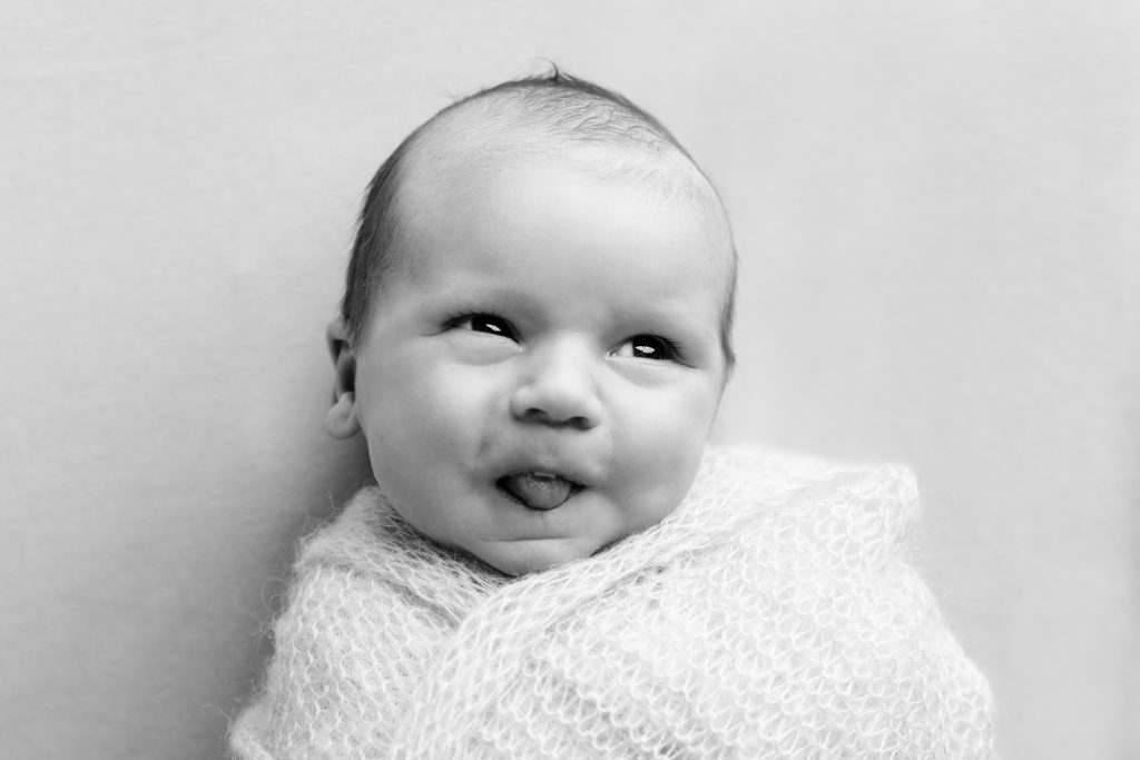 Awake newborn baby wrapped in white making a playful expression in black and white