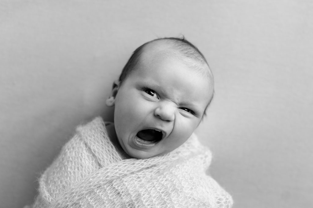 Newborn baby wrapped in white yawning during a black-and-white portrait