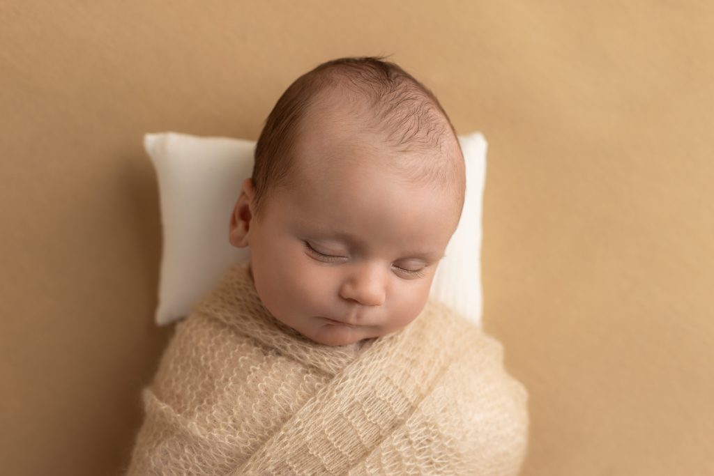 Sleeping newborn swaddled in a soft beige wrap on a warm neutral background