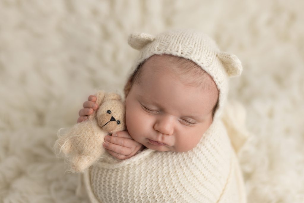 Sleeping newborn wrapped in cream with a knit bear bonnet and tiny teddy bear