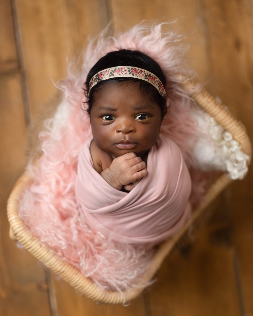 Awake newborn baby wrapped in pink and resting in a woven basket during a portrait session
