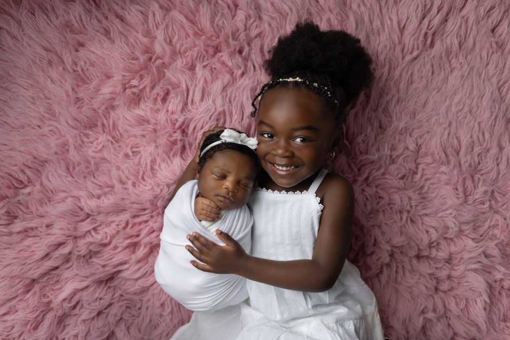 Older sister smiling while holding her sleeping newborn sibling on a soft pink blanket