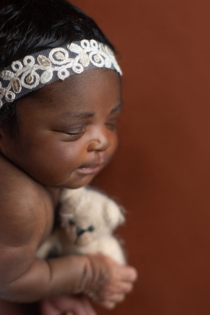 Close-up portrait of a sleeping newborn baby wearing a delicate lace headband and holding a small teddy bear