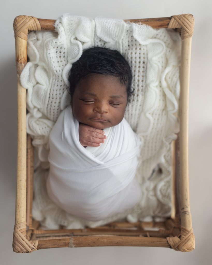 Newborn baby peacefully sleeping while swaddled in white and posed in a woven basket