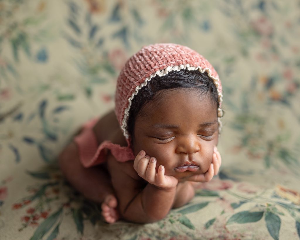 Sleeping newborn baby wearing a knit bonnet and resting her chin on her hands on a floral backdrop