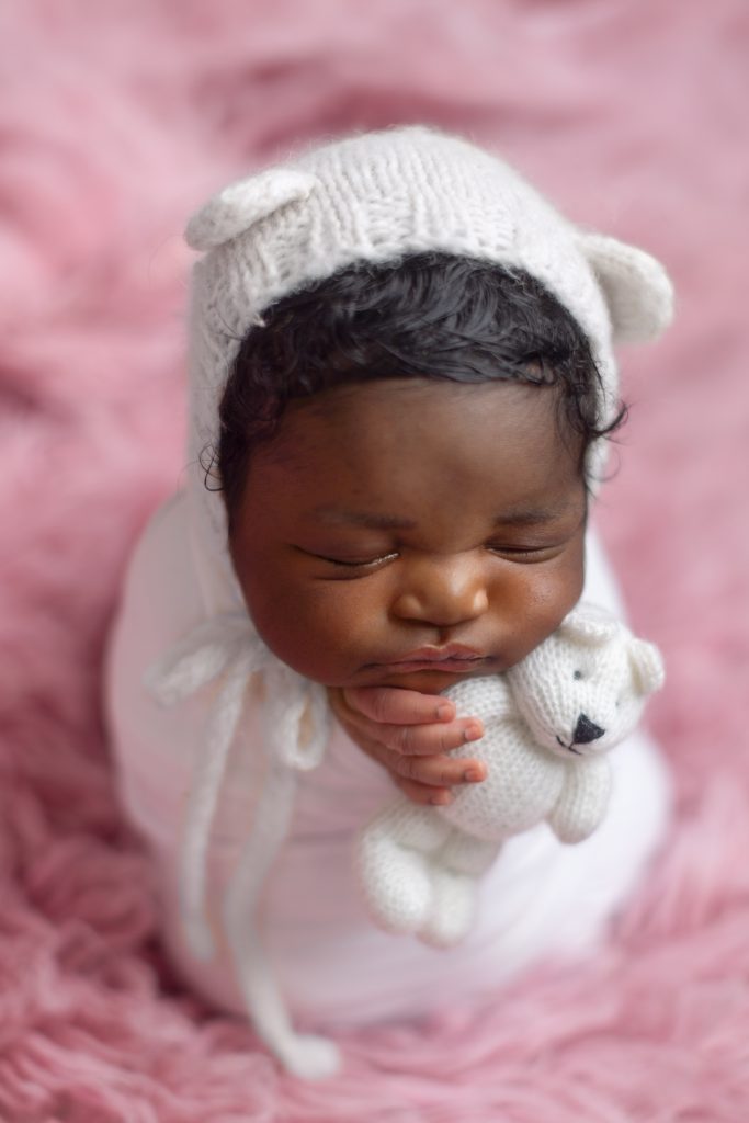 Sleeping newborn baby wearing a white knit hat and cuddling a small teddy bear on a pink blanket