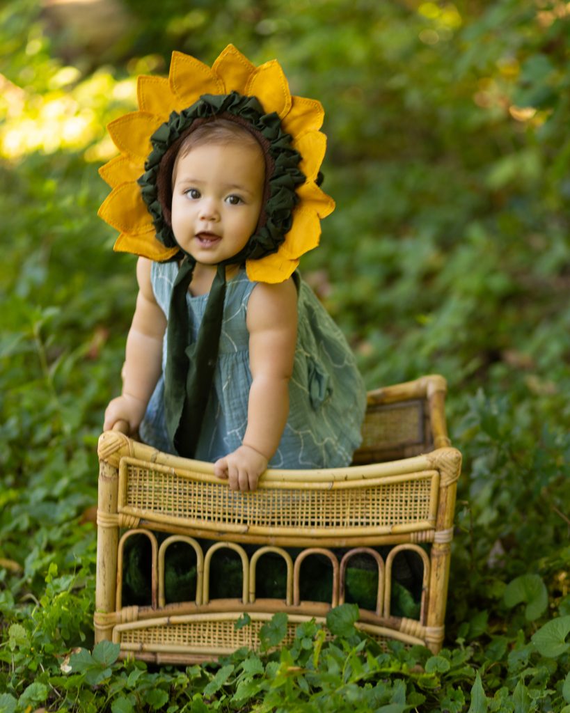 Baby wearing a sunflower bonnet and standing at a wicker chair during an outdoor milestone portrait session.