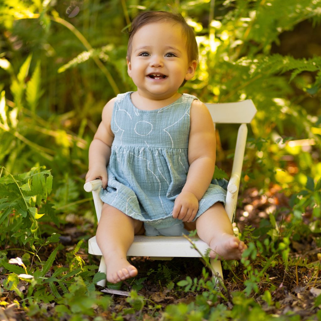 Smiling baby sitting on a small white chair outdoors during a milestone photography session.