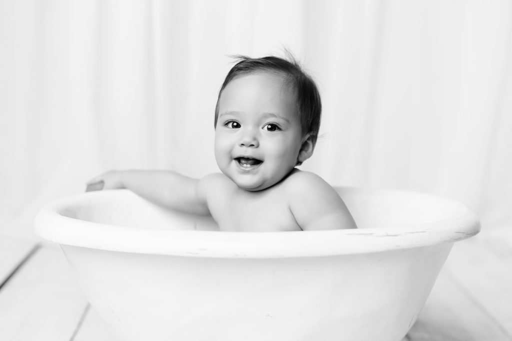 Smiling baby sitting in a white tub during a black-and-white milestone portrait session.