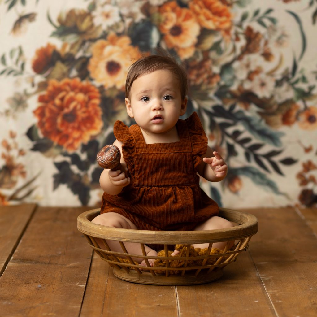 Baby sitting in a wicker basket in a rust-colored outfit during a studio milestone portrait session.