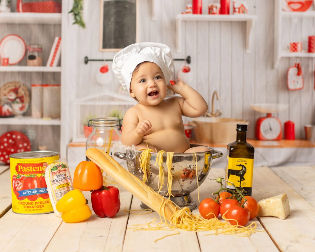 Laughing baby wearing a chef hat and sitting in a colander surrounded by pasta and vegetables.