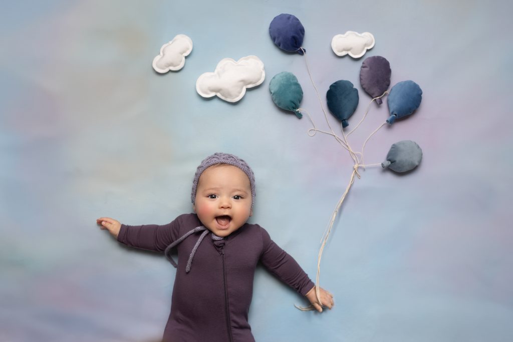 Smiling baby lying beneath soft fabric balloons and clouds during a creative milestone photography session.
