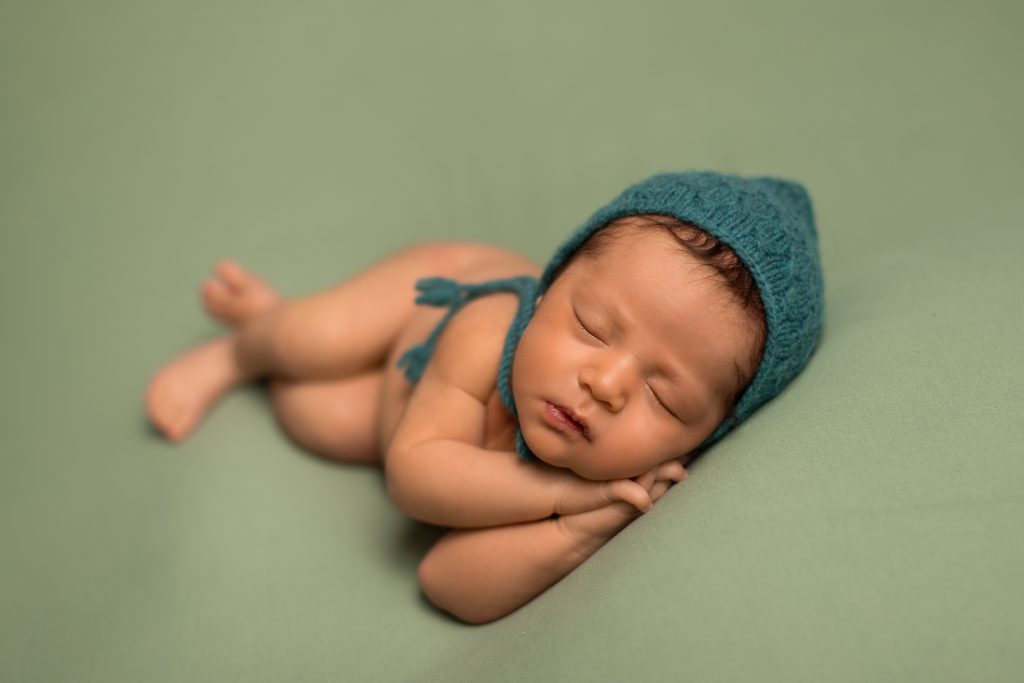 Sleeping newborn wearing a teal knit bonnet on a soft green studio backdrop.