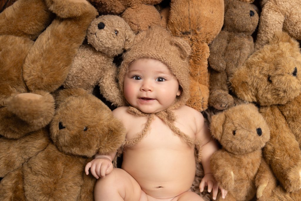 Baby wearing a knitted bear bonnet surrounded by teddy bears during a first-year portrait session.