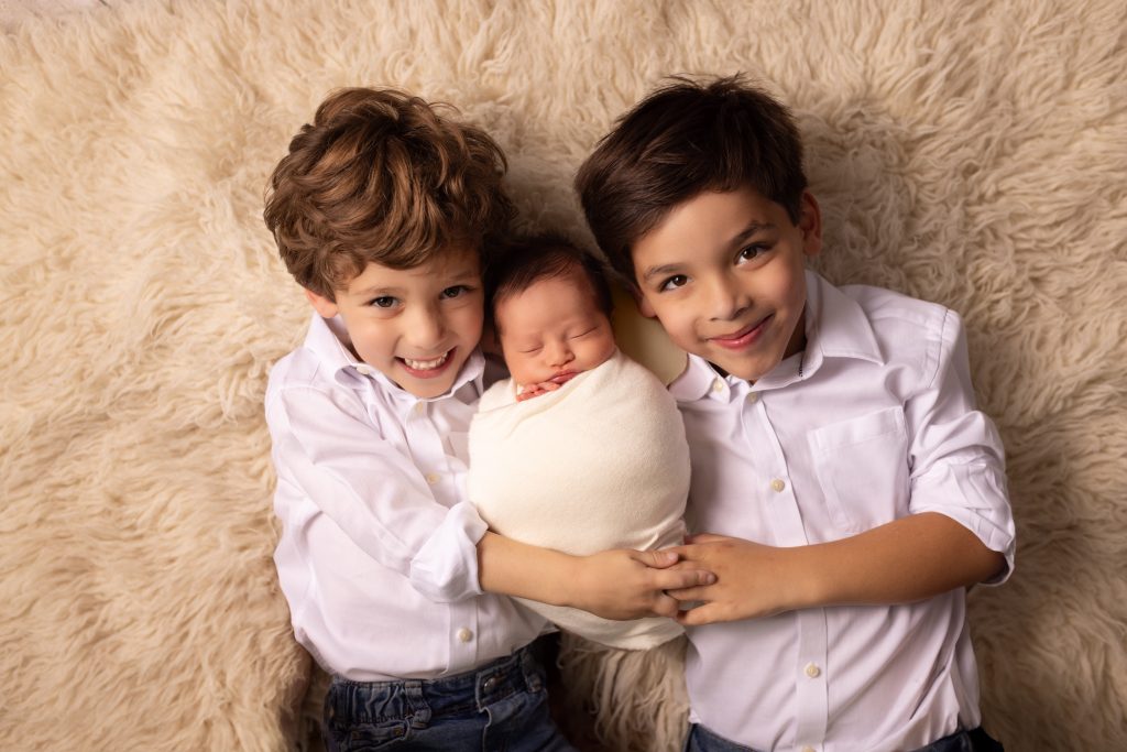Two older brothers cuddling their swaddled newborn sibling during a newborn photography session.