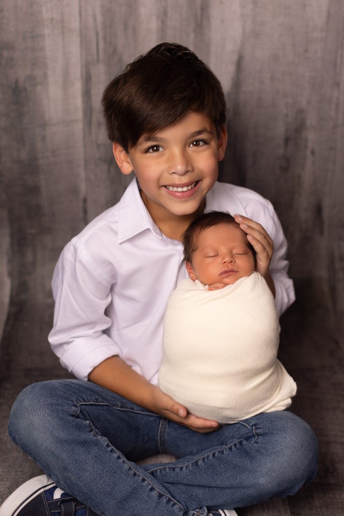 Older brother smiling while holding his swaddled newborn sibling during a studio newborn session.