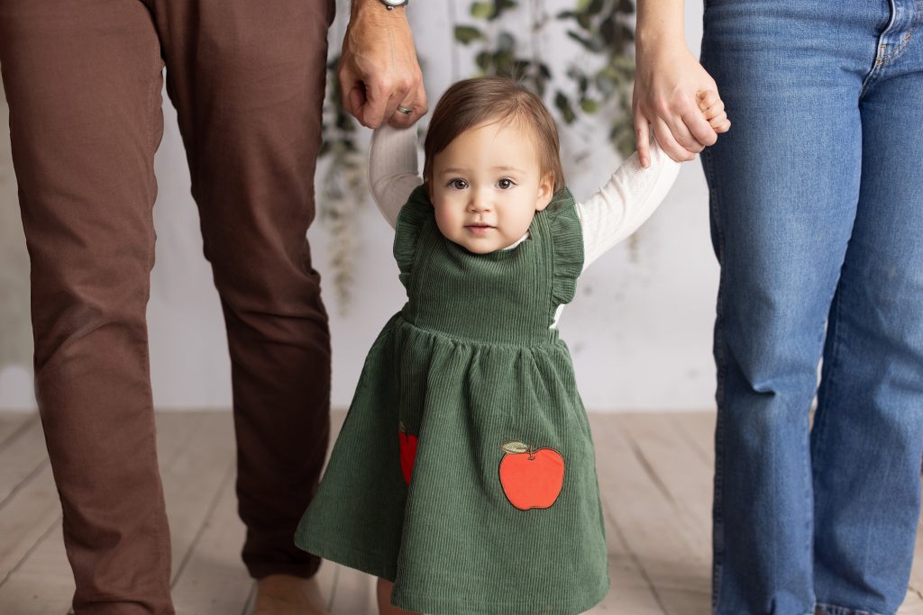 One-year-old baby standing between her parents and holding their hands during a milestone photography session.
