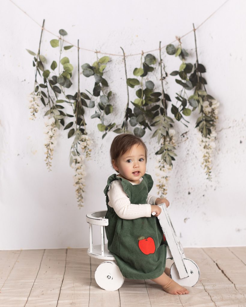 One-year-old child standing beside a white wooden tricycle during a first birthday milestone portrait session.