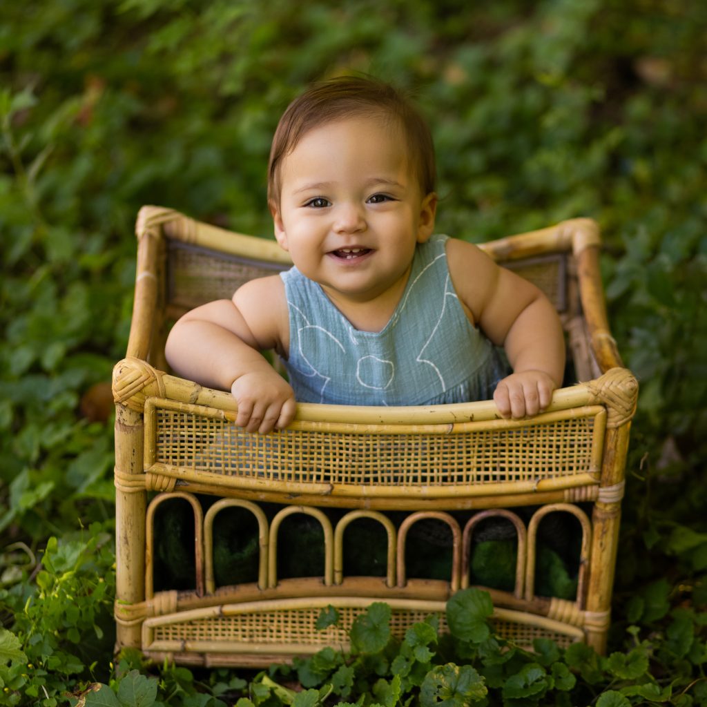 Smiling baby sitting in a vintage wicker chair outdoors during a first-year photography session.