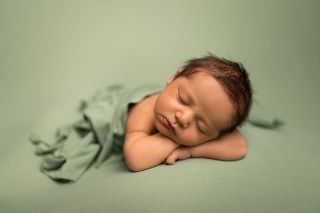 Sleeping newborn resting on folded arms and wrapped in soft green fabric during a studio portrait session.